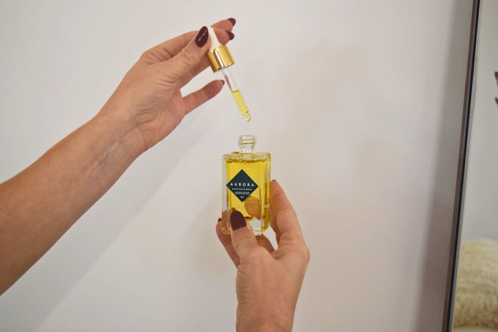 Hands holding and dispensing AURORA saffron-infused oil from a dropper bottle.